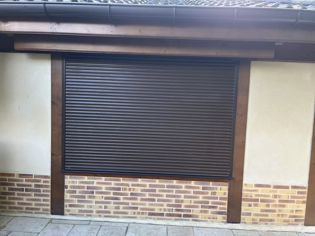 Volet roulant marron foncé installé sur une large fenêtre de maison en briques